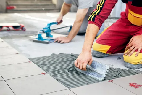 Two tiling professionals from YAM Home Renovation working on a floor tile installation Barrie project; one worker spreads mortar with a trowel while the second worker uses a manual tile cutter.