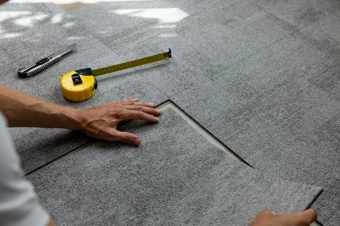 Close-up of hands installing gray modular carpet tiles using a measuring tape and utility knife nearby. YAM Home Renovation provides professional flooring solutions alongside tile installation Barrie.