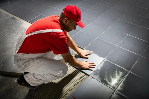 A tiler wearing a red shirt and cap precisely laying down large, square, glossy black floor tiles onto a bed of mortar during a modern tile installation Barrie project by YAM Home Renovation.