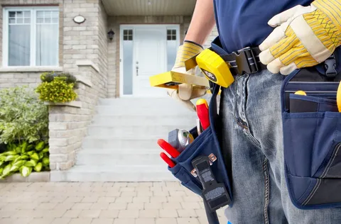 Mid-section view of a skilled contractor holding a level and wearing a tool belt, standing in front of a beautifully renovated stone house entrance. Expert service from YAM Home Renovation, trusted Contractors in Barrie Ontario.